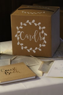 A cardboard box labeled 'Cards' is placed on a table. The box is decorated with a white floral wreath design. Surrounding the box are scattered pages from books and a guest book, which is closed and sits nearby.