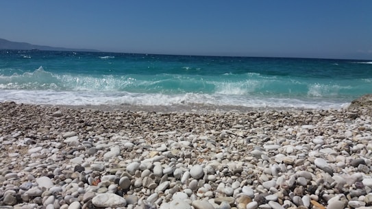 The image depicts a tranquil beach scene with a pebble-covered shoreline leading to clear turquoise waters. Gentle waves are seen lapping against the shore, with the horizon line visible under a clear blue sky.