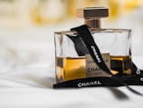 A glass perfume bottle filled with amber-colored liquid, adorned with a black ribbon displaying the brand name. The label indicates it's Gabielle by Chanel, with a sleek, square-shaped design.