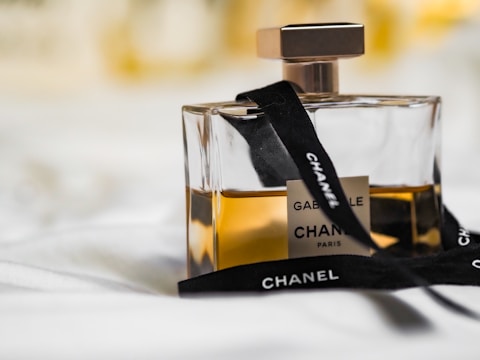 A glass perfume bottle filled with amber-colored liquid, adorned with a black ribbon displaying the brand name. The label indicates it's Gabielle by Chanel, with a sleek, square-shaped design.