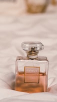 A perfume bottle with a rectangular shape and a transparent outer layer showcasing a pale amber liquid inside. It has a label with gold accents and is set against a soft, white fabric backdrop.