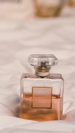 A perfume bottle with a rectangular shape and a transparent outer layer showcasing a pale amber liquid inside. It has a label with gold accents and is set against a soft, white fabric backdrop.