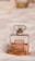 A perfume bottle with a rectangular shape and a transparent outer layer showcasing a pale amber liquid inside. It has a label with gold accents and is set against a soft, white fabric backdrop.