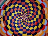 A vibrant and symmetric pattern of concentric circles composed of alternating blue, yellow, orange, and red squares. The design is reminiscent of a kaleidoscope, with lines radiating from the center creating a sense of depth.