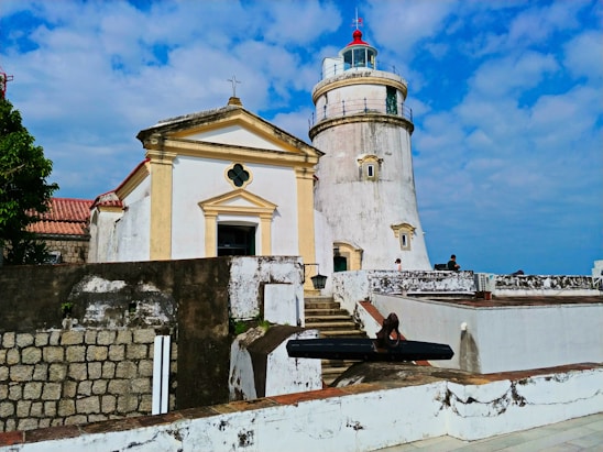 A historical site featuring a white and beige lighthouse adjacent to a small chapel with a pitched roof and a cross on top. The buildings are weathered, indicating age, with some people visible on the structure's roof in a casual setting. The sky is bright blue with scattered clouds.