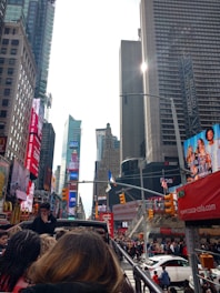 A bustling cityscape with tall skyscrapers, large billboards, and a crowd of people. Bright advertisements and signs fill the scene, capturing the vibrant and busy energy of an urban center. The sky is bright and partly cloudy.