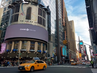 A bustling urban street scene featuring tall buildings with large digital billboards displaying advertisements. A prominent advertisement for a Chase bank is visible. Yellow taxis are driving on the street, and pedestrians are walking on the sidewalks. The sky is partly cloudy, and the atmosphere is lively.