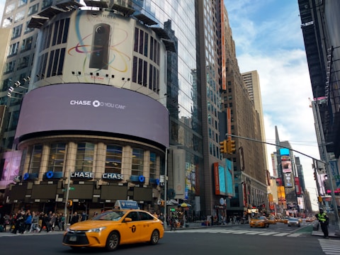 A bustling urban street scene featuring tall buildings with large digital billboards displaying advertisements. A prominent advertisement for a Chase bank is visible. Yellow taxis are driving on the street, and pedestrians are walking on the sidewalks. The sky is partly cloudy, and the atmosphere is lively.