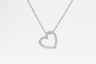 A heart-shaped pendant encrusted with small diamonds is suspended from a delicate chain. The chain appears to be made of tiny, round metal beads, and the pendant sparkles against a white background.