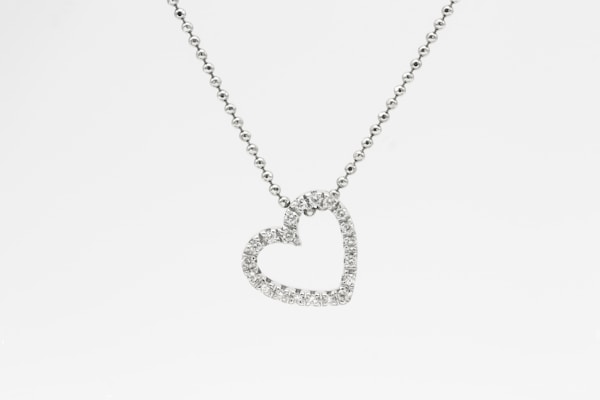A heart-shaped pendant encrusted with small diamonds is suspended from a delicate chain. The chain appears to be made of tiny, round metal beads, and the pendant sparkles against a white background.