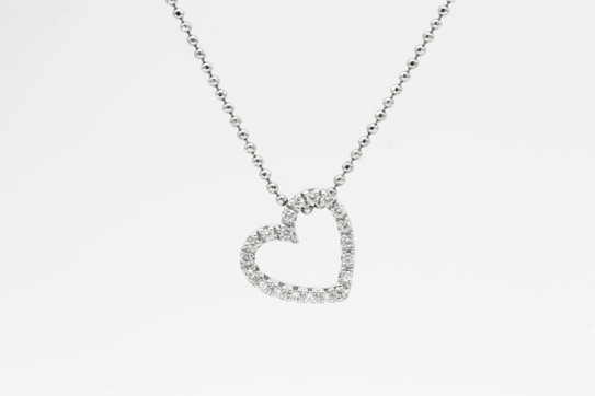 A heart-shaped pendant encrusted with small diamonds is suspended from a delicate chain. The chain appears to be made of tiny, round metal beads, and the pendant sparkles against a white background.