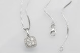 A silver necklace with a square pendant encrusted with multiple small diamonds is laid out on a flat surface. The chain is delicate and features a lobster clasp closure. The overall look is elegant and sophisticated.