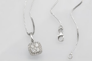 A silver necklace with a square pendant encrusted with multiple small diamonds is laid out on a flat surface. The chain is delicate and features a lobster clasp closure. The overall look is elegant and sophisticated.