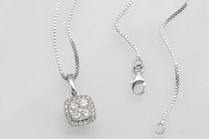 A silver necklace with a square pendant encrusted with multiple small diamonds is laid out on a flat surface. The chain is delicate and features a lobster clasp closure. The overall look is elegant and sophisticated.