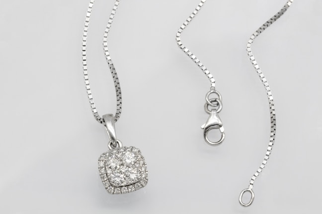 A silver necklace with a square pendant encrusted with multiple small diamonds is laid out on a flat surface. The chain is delicate and features a lobster clasp closure. The overall look is elegant and sophisticated.