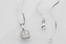 A silver necklace with a square pendant encrusted with multiple small diamonds is laid out on a flat surface. The chain is delicate and features a lobster clasp closure. The overall look is elegant and sophisticated.