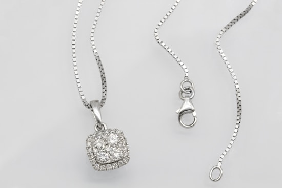 A silver necklace with a square pendant encrusted with multiple small diamonds is laid out on a flat surface. The chain is delicate and features a lobster clasp closure. The overall look is elegant and sophisticated.