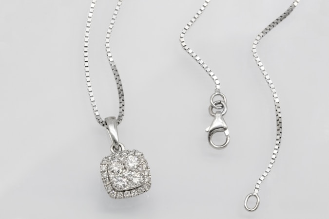 A silver necklace with a square pendant encrusted with multiple small diamonds is laid out on a flat surface. The chain is delicate and features a lobster clasp closure. The overall look is elegant and sophisticated.
