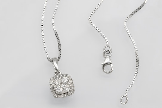 A silver necklace with a square pendant encrusted with multiple small diamonds is laid out on a flat surface. The chain is delicate and features a lobster clasp closure. The overall look is elegant and sophisticated.