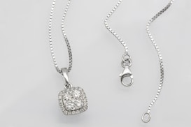 A silver necklace with a square pendant encrusted with multiple small diamonds is laid out on a flat surface. The chain is delicate and features a lobster clasp closure. The overall look is elegant and sophisticated.