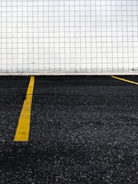 A section of a parking lot with yellow lines on the asphalt indicating parking spaces. The background consists of a wall covered in white tiles arranged in a grid pattern.