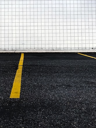 A section of a parking lot with yellow lines on the asphalt indicating parking spaces. The background consists of a wall covered in white tiles arranged in a grid pattern.