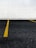 A section of a parking lot with yellow lines on the asphalt indicating parking spaces. The background consists of a wall covered in white tiles arranged in a grid pattern.