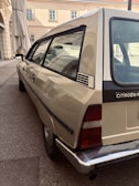 A classic Citroën car is parked on a paved street near a building with beige walls and multiple windows. The vehicle has a distinctive retro design, and the scene is calm with muted earth tones.
