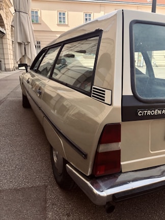 A classic Citroën car is parked on a paved street near a building with beige walls and multiple windows. The vehicle has a distinctive retro design, and the scene is calm with muted earth tones.