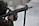 A person in camouflage uniform aiming a rifle with precision. The individual's head is slightly turned to the left, wearing protective ear gear. The background is blurred, adding focus to the subject.