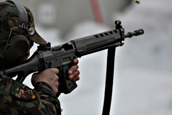 A person in camouflage uniform aiming a rifle with precision. The individual's head is slightly turned to the left, wearing protective ear gear. The background is blurred, adding focus to the subject.