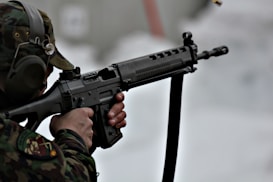 A person in camouflage uniform aiming a rifle with precision. The individual's head is slightly turned to the left, wearing protective ear gear. The background is blurred, adding focus to the subject.