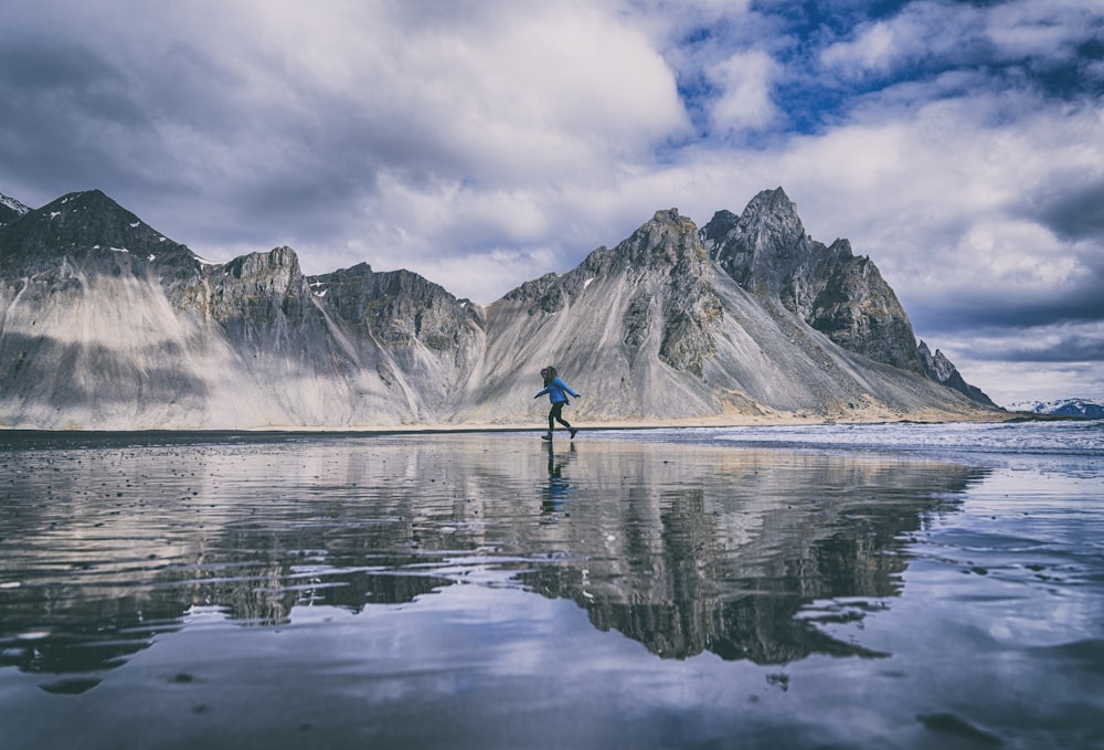 person in blue shirt and black pants standing on blue kayak on lake near mountain under