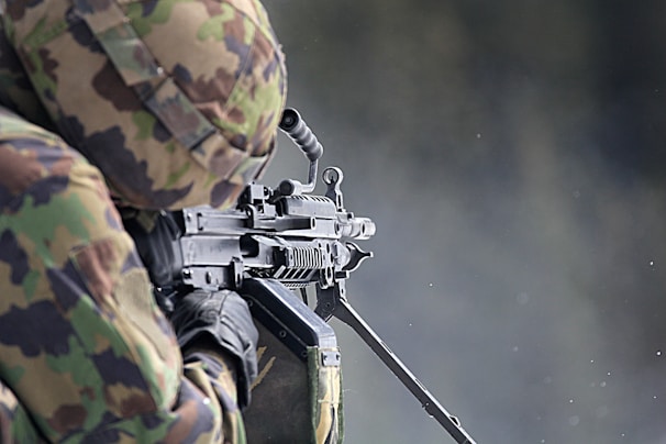 A person wearing camouflage clothing is handling a heavy machine gun, with focus on the weapon's intricate details. The background is blurred, creating a contrast that highlights the gun and the soldier's gear.