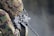 A person wearing camouflage clothing is handling a heavy machine gun, with focus on the weapon's intricate details. The background is blurred, creating a contrast that highlights the gun and the soldier's gear.