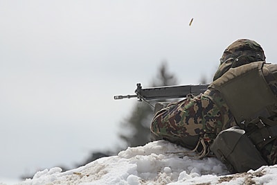 A hunter in camo gear crouching quietly near a frozen pond surrounded by bare winter trees.
