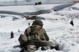 A soldier in camouflage uniform lies prone on a snowy landscape during a military exercise. The scene is set in a wintry environment with snow-covered ground and targets in the distance. The soldier is equipped with a helmet and a backpack, and appears to be engaged in a training activity.
