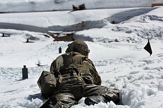 A soldier in camouflage uniform lies prone on a snowy landscape during a military exercise. The scene is set in a wintry environment with snow-covered ground and targets in the distance. The soldier is equipped with a helmet and a backpack, and appears to be engaged in a training activity.