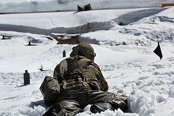 A soldier in camouflage uniform lies prone on a snowy landscape during a military exercise. The scene is set in a wintry environment with snow-covered ground and targets in the distance. The soldier is equipped with a helmet and a backpack, and appears to be engaged in a training activity.