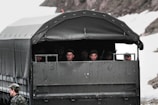 A military vehicle with a canvas-covered back carries several uniformed soldiers, who are sitting and looking outward. The vehicle is parked on a snowy terrain, and one soldier is standing outside the vehicle wearing a camouflage uniform and cap.