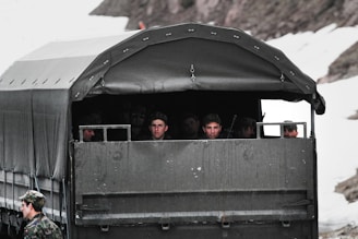A military vehicle with a canvas-covered back carries several uniformed soldiers, who are sitting and looking outward. The vehicle is parked on a snowy terrain, and one soldier is standing outside the vehicle wearing a camouflage uniform and cap.