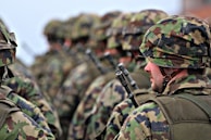 Soldiers in camouflage uniforms and helmets standing in formation with weapons visible. The focus is on the soldier in the foreground wearing a green and brown patterned helmet.