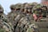 Soldiers in camouflage uniforms and helmets standing in formation with weapons visible. The focus is on the soldier in the foreground wearing a green and brown patterned helmet.