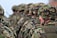 Soldiers in camouflage uniforms and helmets standing in formation with weapons visible. The focus is on the soldier in the foreground wearing a green and brown patterned helmet.