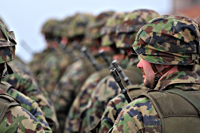 Soldiers in camouflage uniforms and helmets standing in formation with weapons visible. The focus is on the soldier in the foreground wearing a green and brown patterned helmet.