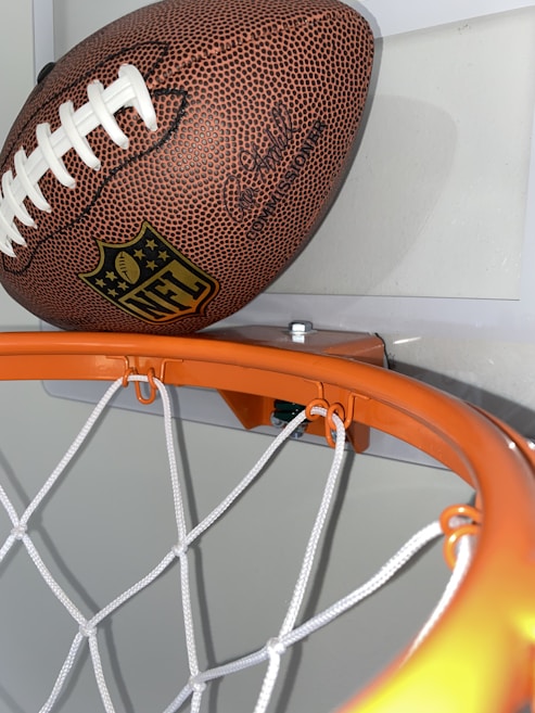 A football is positioned on a basketball hoop, with its distinct textured surface and white laces prominently visible. The hoop's orange rim and white netting stand out against the neutral background.