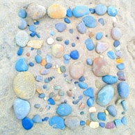 A collection of multicolored pebbles arranged in a rectangular pattern on sandy ground. The stones vary in size and include shades of blue, orange, and grey, creating a visually pleasing composition. The background consists of fine beige sand.