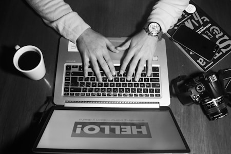 A person wearing a watch is typing on a laptop. A coffee mug, camera, smartphone, and a magazine are situated on the wooden table surrounding the laptop. An inverted tablet displays the word 'HELLO!'. The scene is monochromatic, creating a classic, timeless feel.