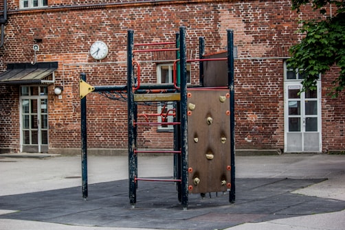 An outdoor playground set next to a brick building features a structure with climbing holds and a ladder. The building has large windows and a clock is mounted on the wall. A canopy above a door is visible, along with a patch of greenery from a tree.