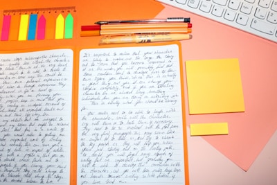 A workspace with an open notebook filled with handwritten notes. Around the notebook, there are colorful sticky notes, a ruler, several pens, and a section of a keyboard visible. The background is a warm orange and pink color scheme.